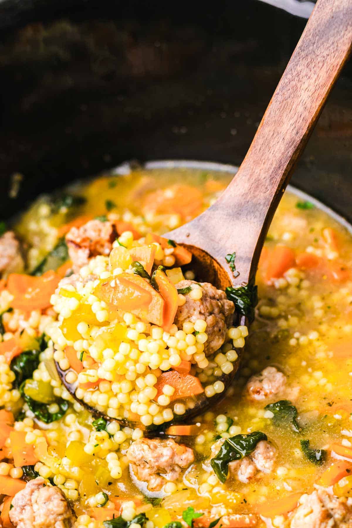 A wooden spoon holds a comforting blend of couscous soup with carrots, greens, and meatballs over a pot filled with savory broth, reminiscent of classic Slow Cooker Italian Wedding Soup.
