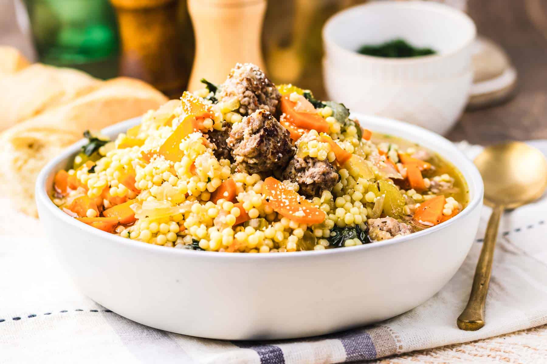 A bowl reminiscent of Slow Cooker Italian Wedding Soup, featuring couscous with meatballs, carrots, celery, and greens, served on a table with bread and a gold spoon.