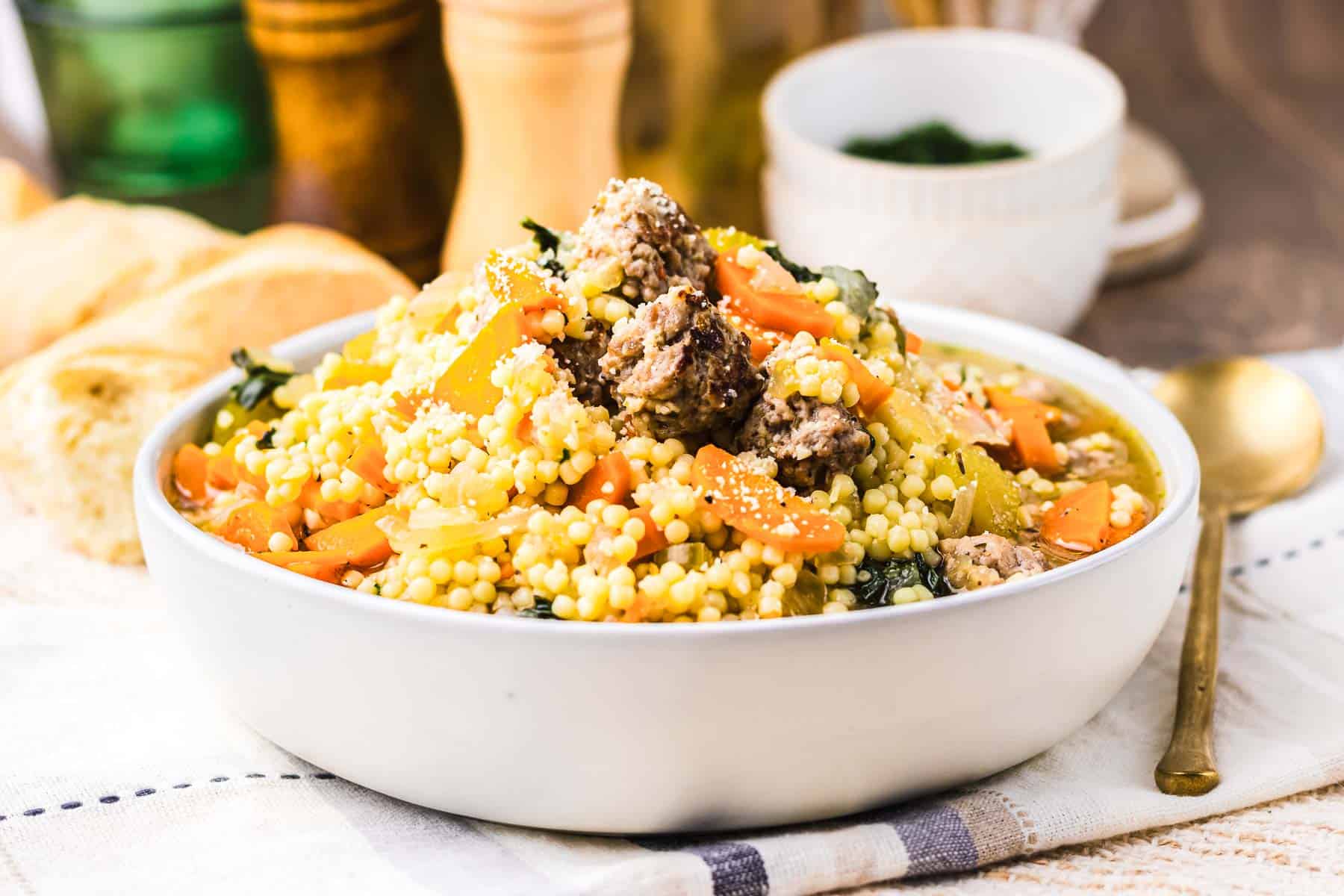 A bowl of couscous with vegetables and meat, reminiscent of the comforting flavors found in Slow Cooker Italian Wedding Soup, is served on a table with bread and a gold spoon nearby.