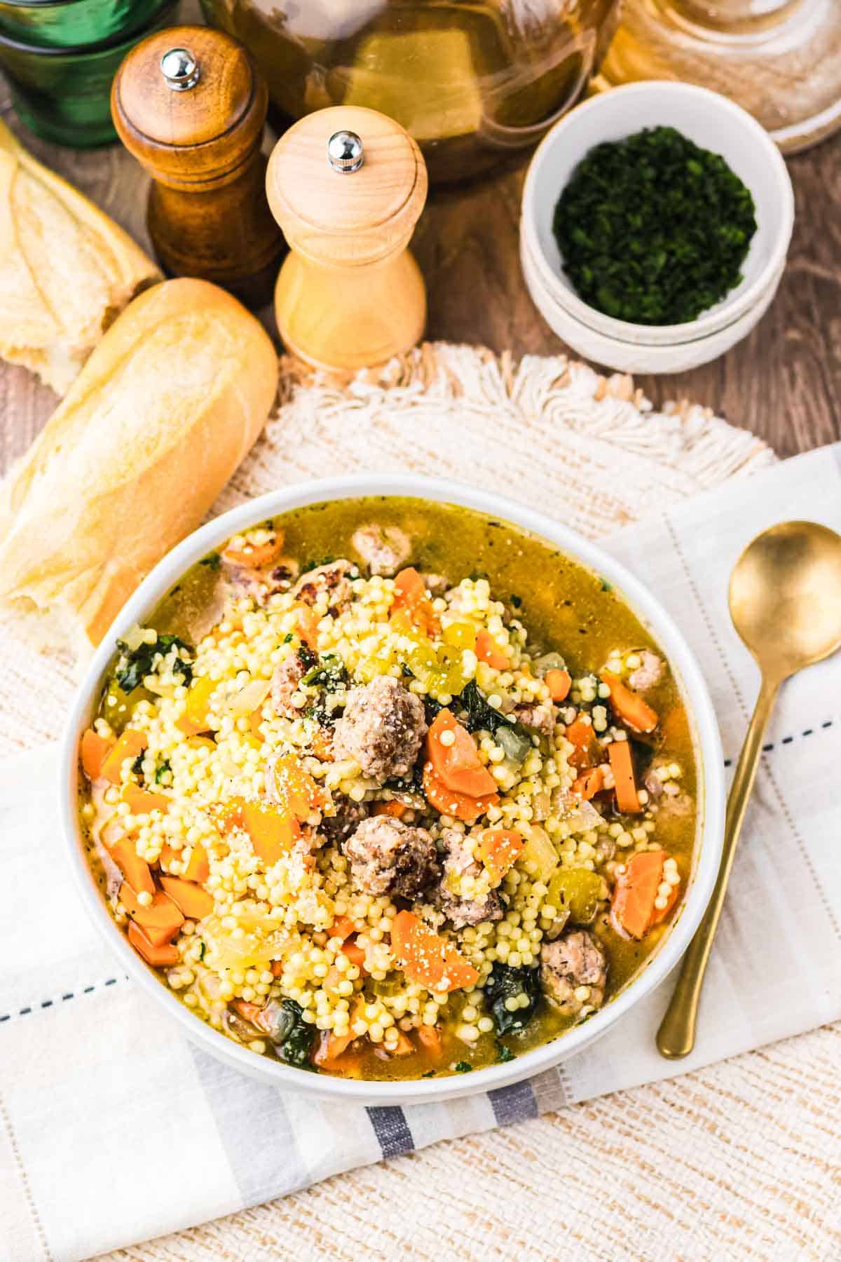 Enjoy a comforting bowl of couscous soup with vegetables and sausage, reminiscent of Slow Cooker Italian Wedding Soup, served beside fresh bread, herbs, and classic salt and pepper shakers.
