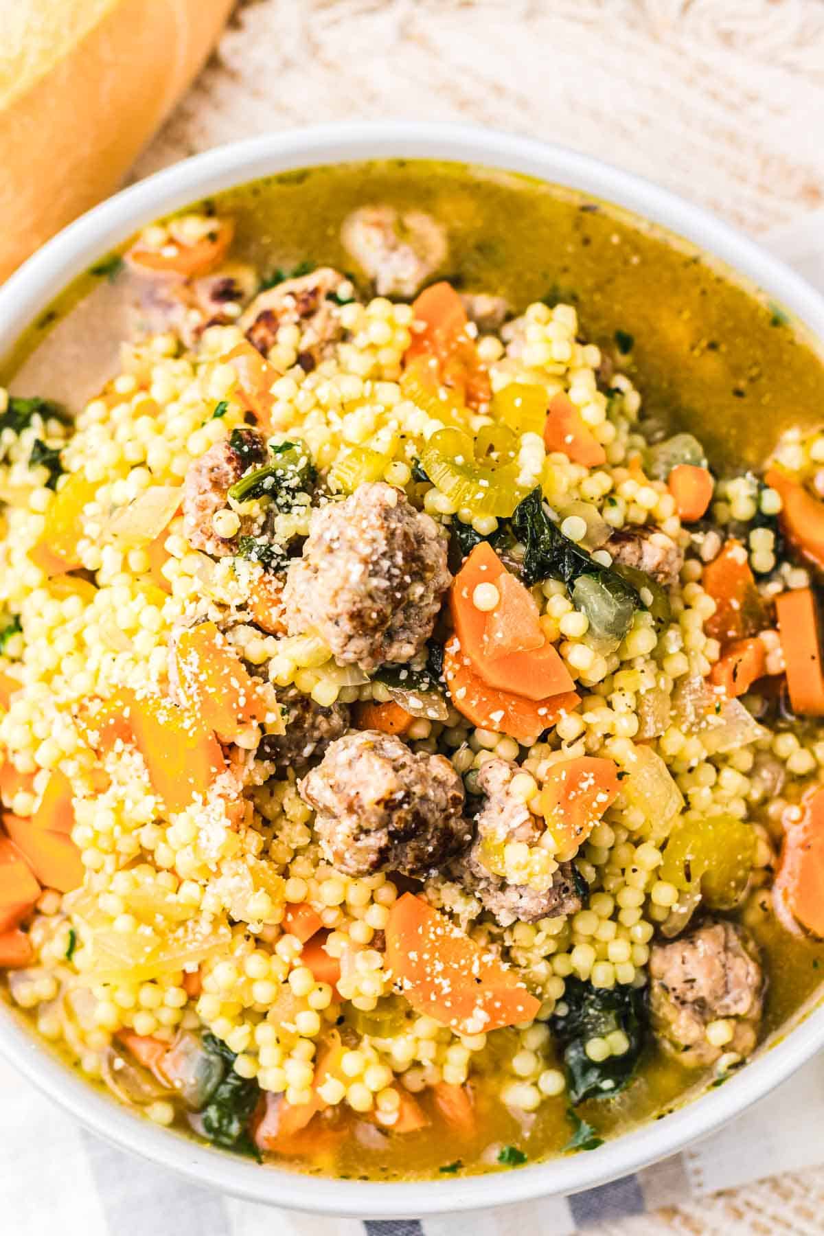Bowl of Slow Cooker Italian Wedding Soup with couscous, meatballs, carrots, celery, and greens on a light surface.