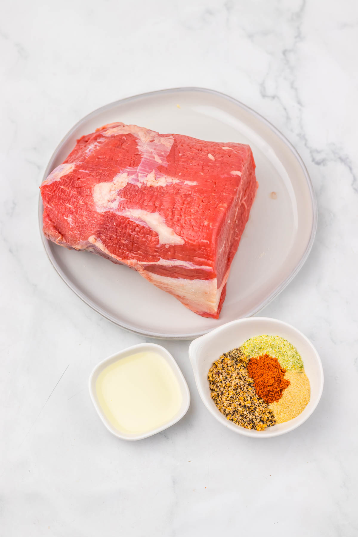 Raw beef on a plate with a small bowl of oil and a bowl of mixed spices on a white marble background—perfectly prepared for making an Air Fryer Eye of Round Roast.
