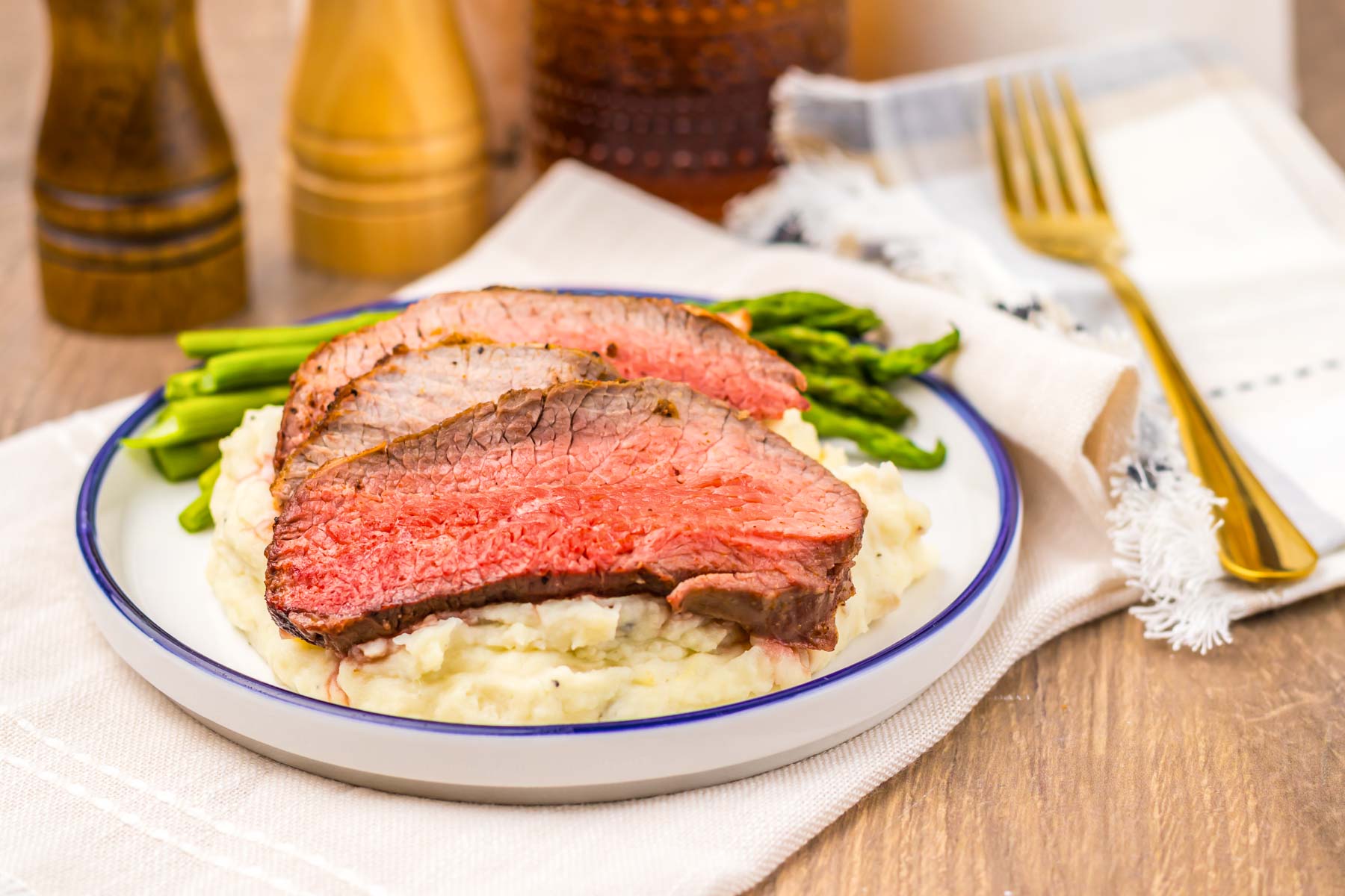 Sliced Air Fryer Eye of Round Roast on mashed potatoes with green beans, served on a white plate with a fork and napkin.