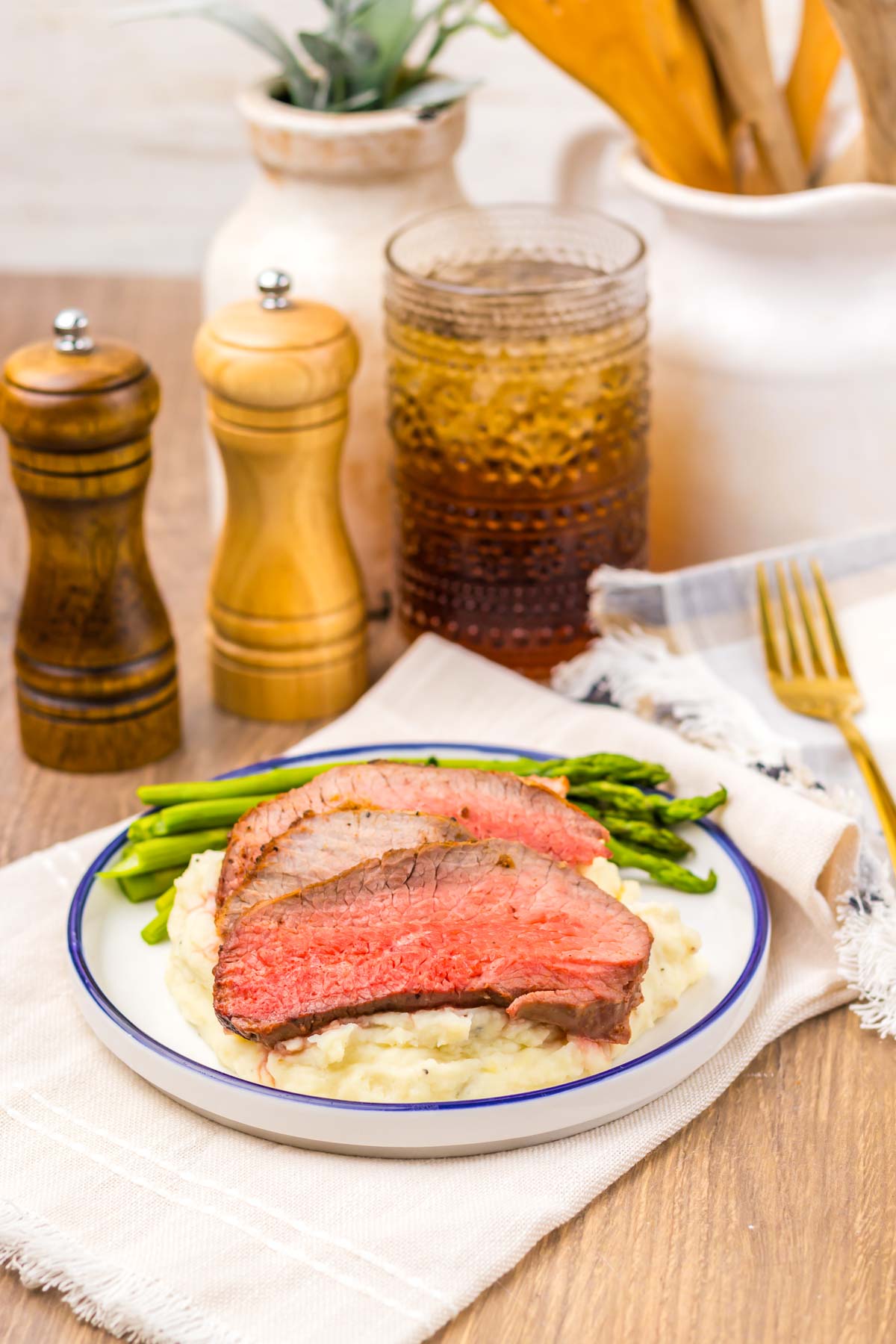 Two slices of tender Air Fryer Eye of Round Roast rest atop creamy mashed potatoes with asparagus, elegantly served on a plate alongside refreshing drinks and condiments.