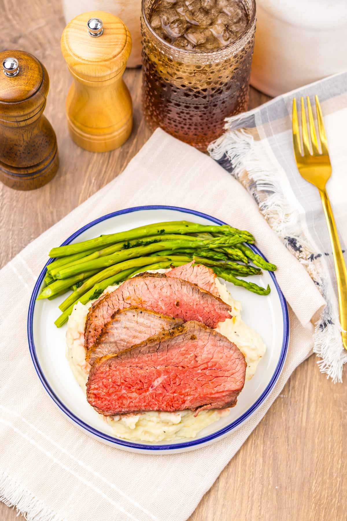 Three slices of Air Fryer Eye of Round Roast, asparagus, and mashed potatoes are elegantly arranged on a plate alongside a drink, napkin, and utensils.