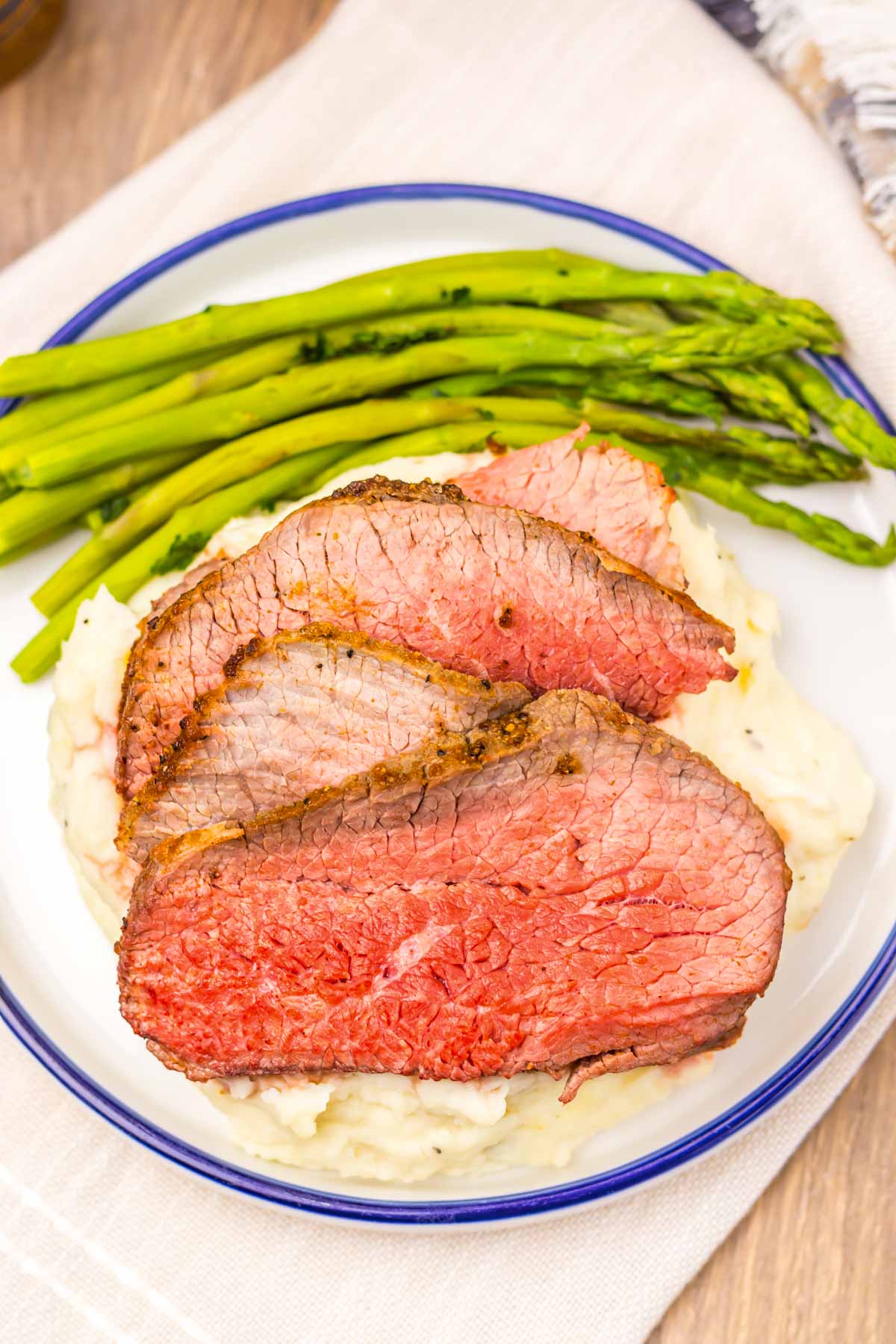 Three slices of Air Fryer Eye of Round Roast rest atop a bed of creamy mashed potatoes, accompanied by tender asparagus spears on a pristine white plate.