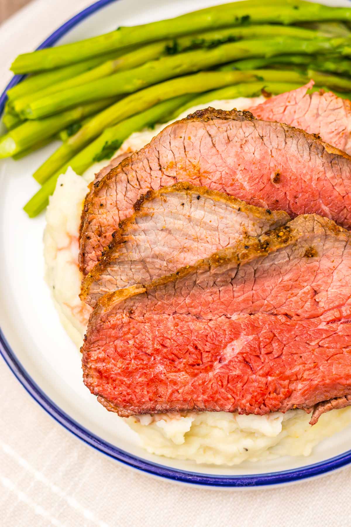 Three slices of air fryer eye of round roast rest atop creamy mashed potatoes, accompanied by crisp asparagus spears on a white plate.