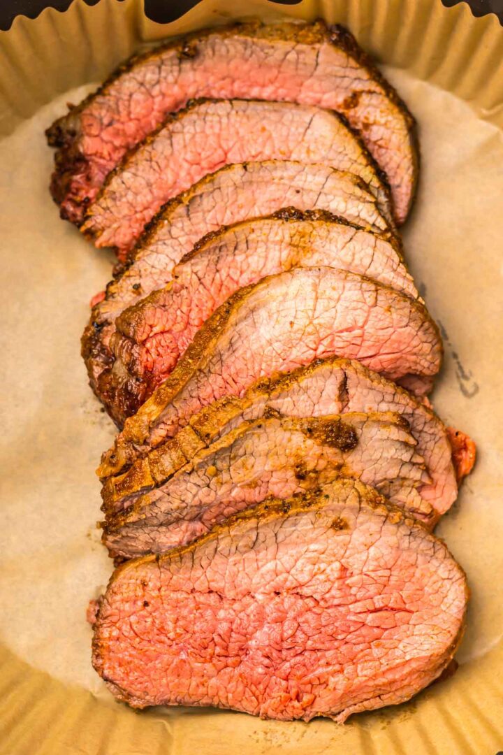 Six slices of medium-rare roast beef, prepared from a perfectly cooked Air Fryer Eye of Round Roast, are arranged on brown parchment paper in a baking dish.