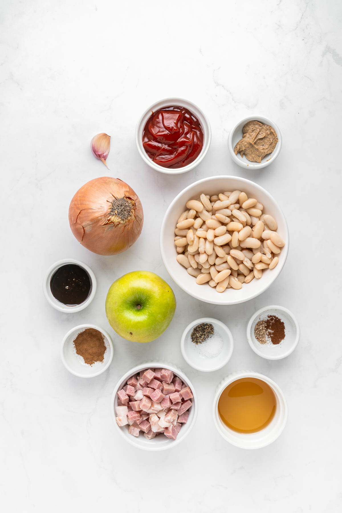 Bowls of beans, apple, onion, ham, ketchup, spices, garlic, and mustard arranged on a white surface.