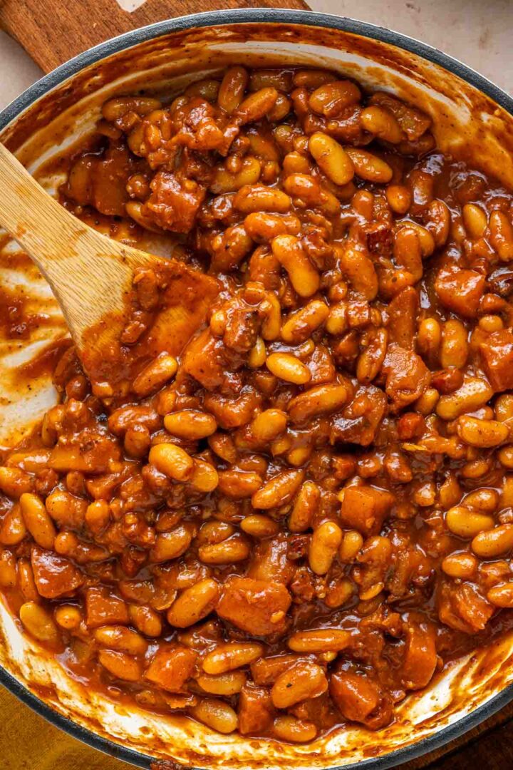 Apple Pie Baked Beans mixed with chunks of meat in a thick sauce, stirred with a wooden spoon in a pot.