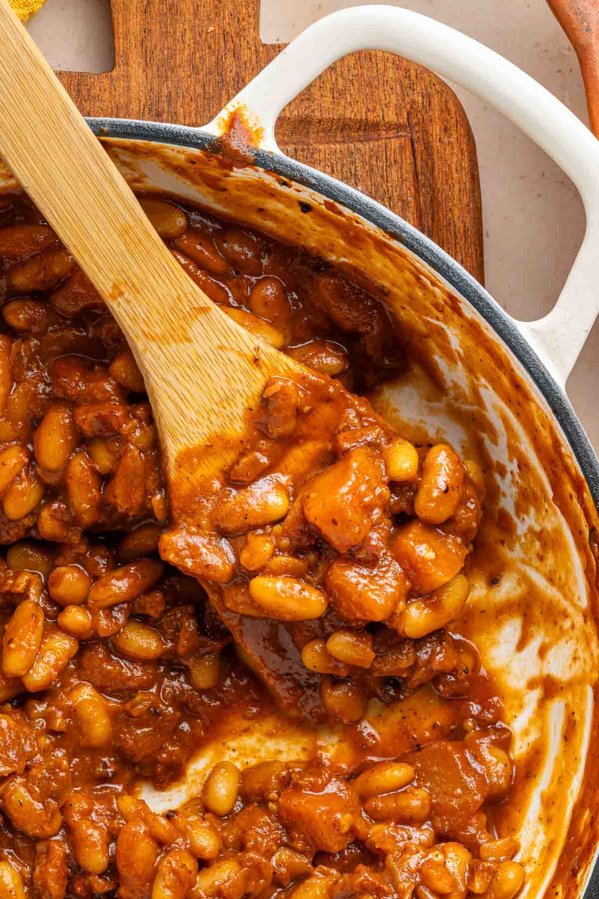 Apple Pie Baked Beans in a white pot with a wooden spoon on a wooden cutting board.