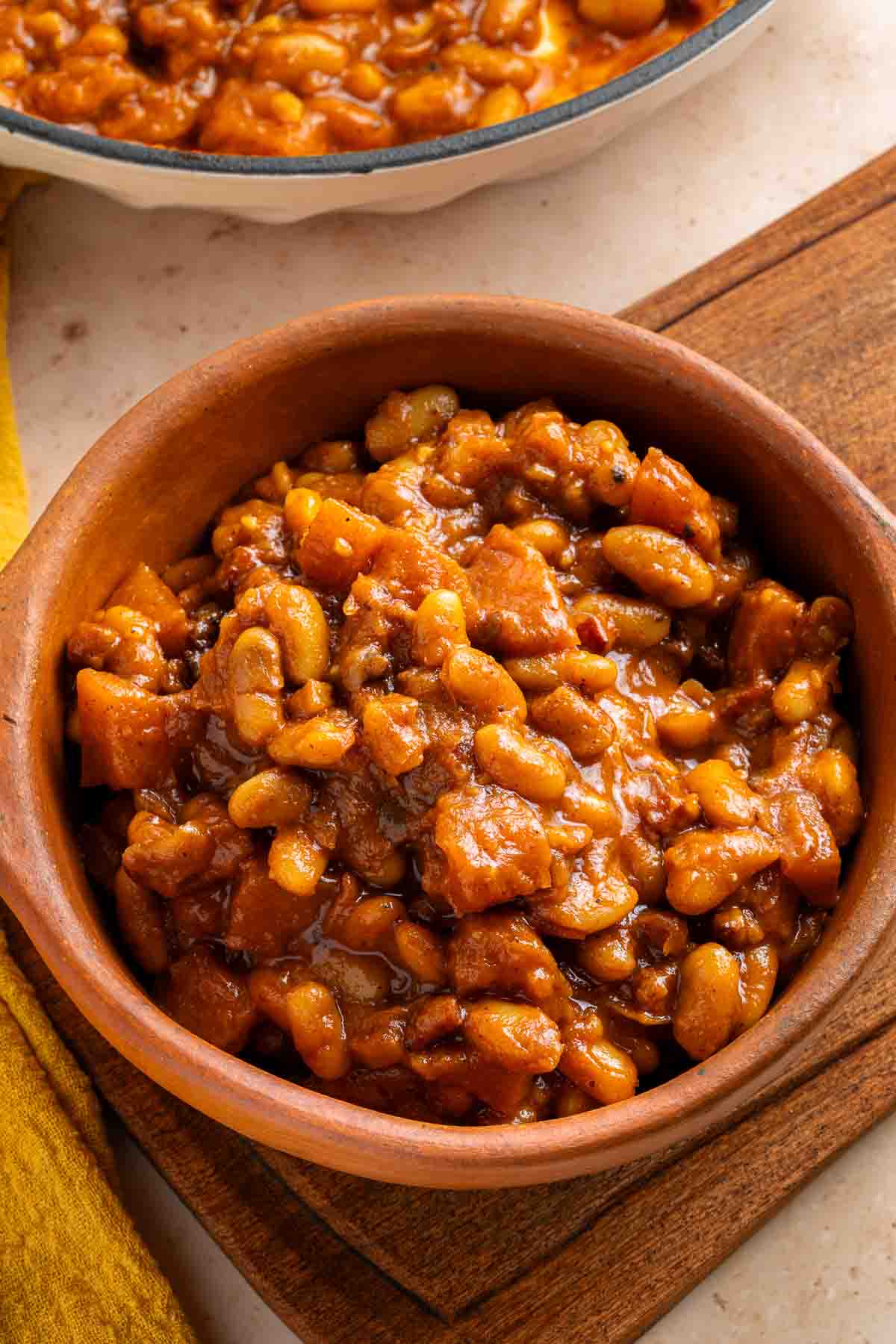 A brown ceramic bowl filled with Apple Pie Baked Beans in a thick, rich sauce sits on a wooden board.