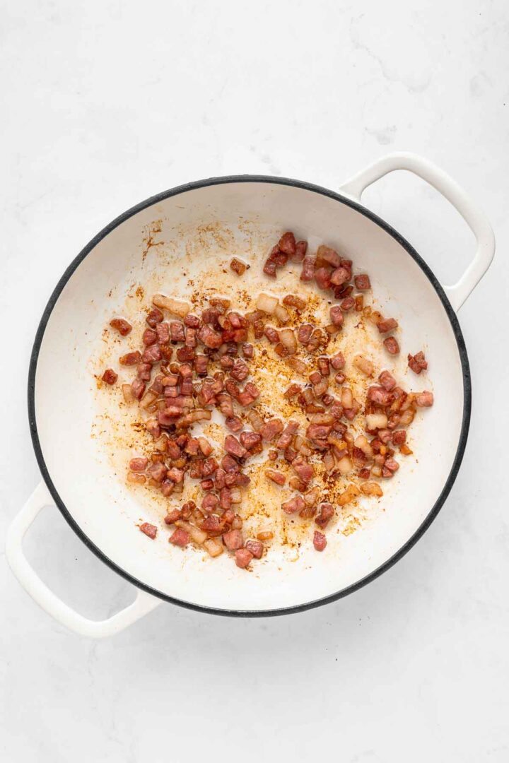 White pot with browned diced pancetta and onions sauteing on the bottom, perfect as a savory base for Apple Pie Baked Beans, on a white surface.