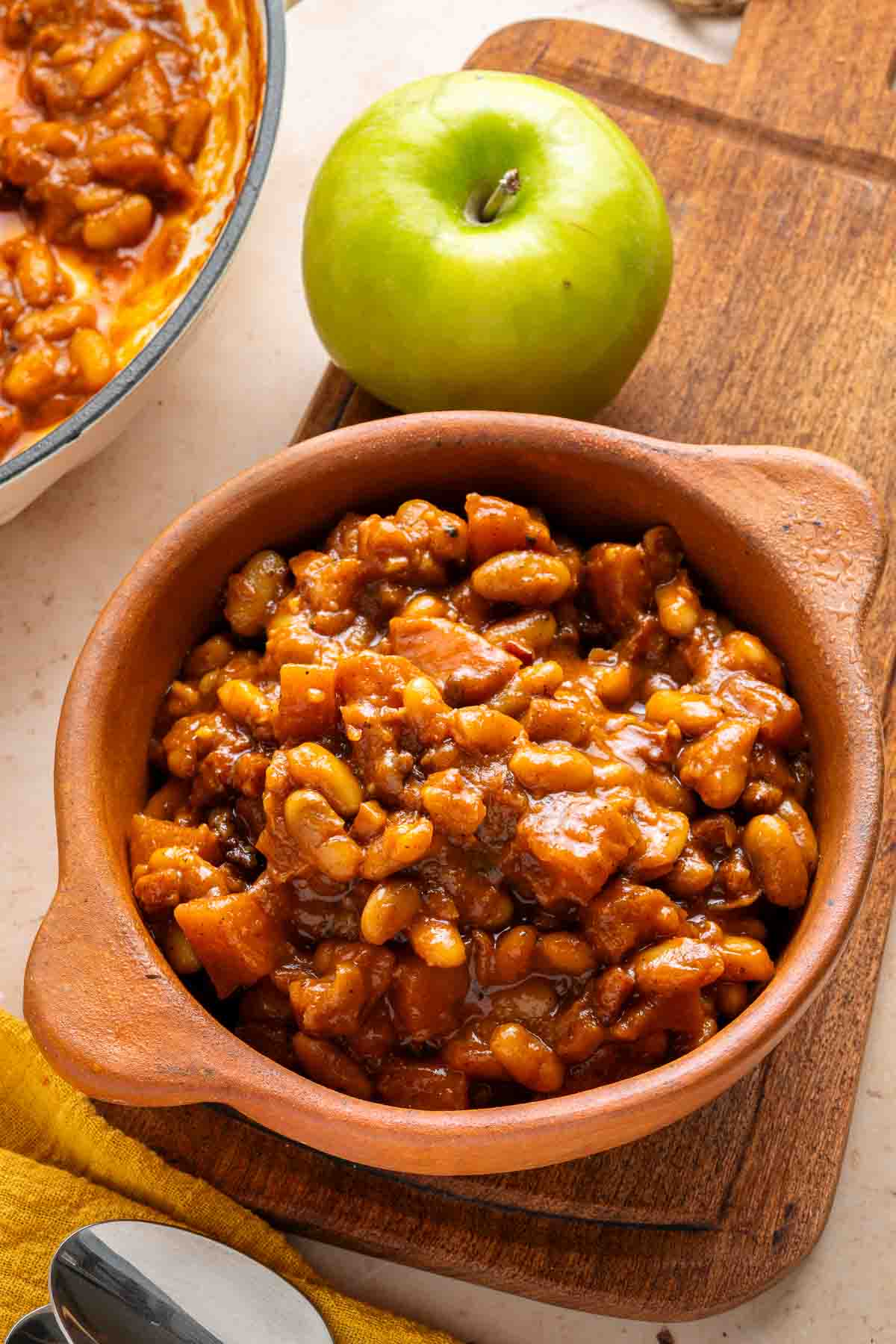 A brown bowl of Apple Pie Baked Beans sits on a wooden board next to a green apple and a pan of beans.