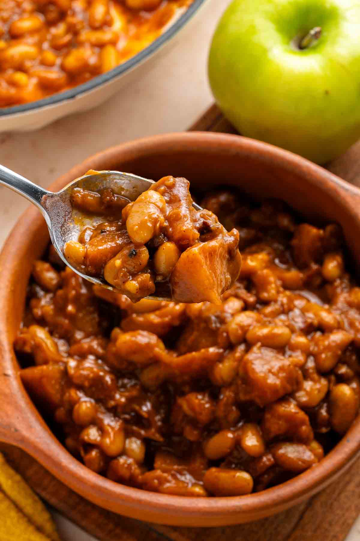 Spoonful of Apple Pie Baked Beans with chunks of meat in a brown bowl, green apple nearby.