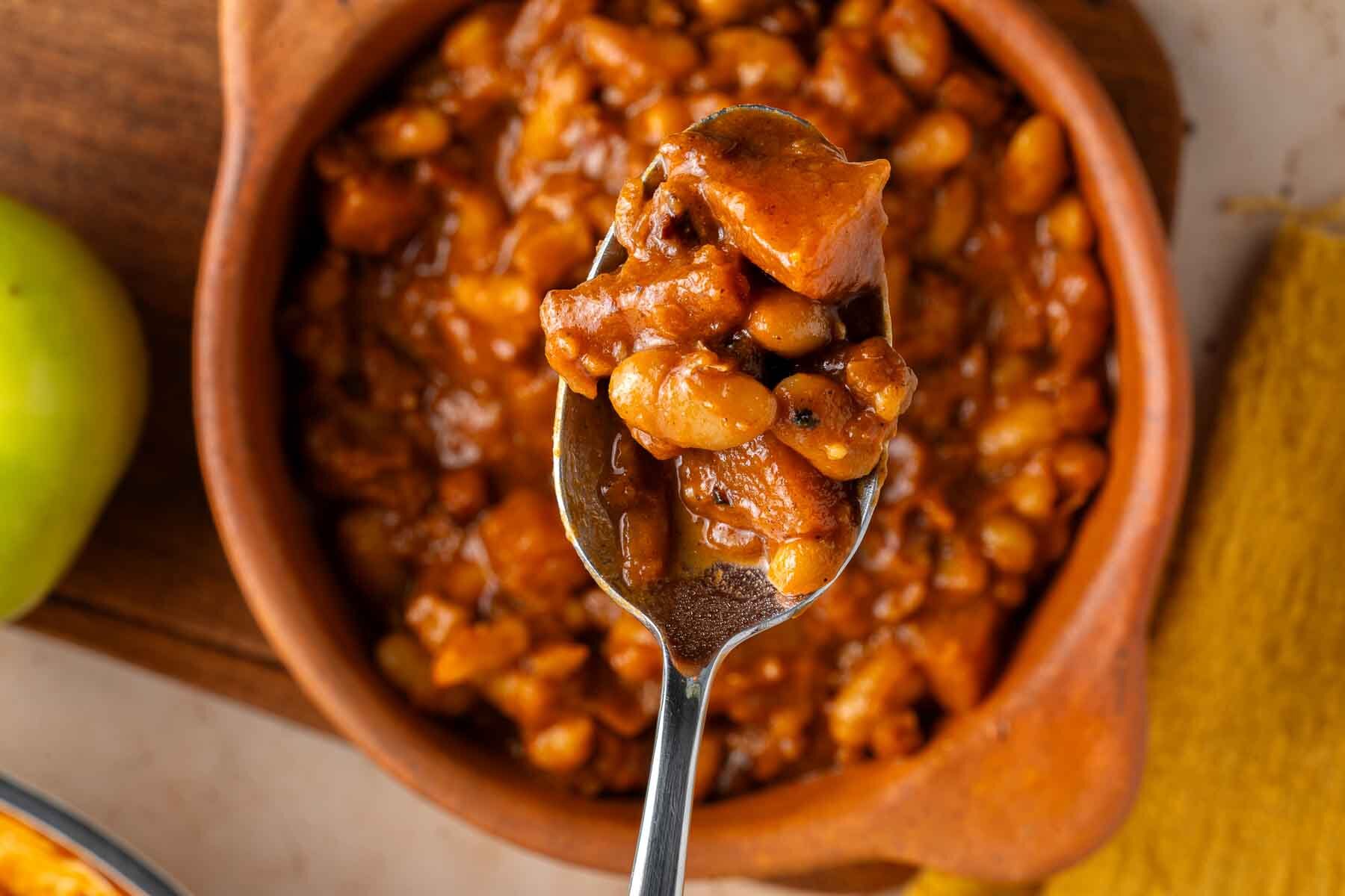 A spoonful of Apple Pie Baked Beans with chunks of sausage held over a wooden bowl filled with baked beans.