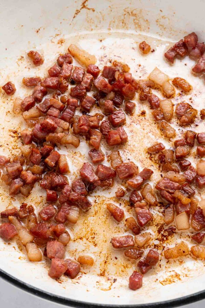 Diced bacon pieces sizzling and browning in a white pan with visible rendered fat, ready to add a savory touch to Apple Pie Baked Beans.