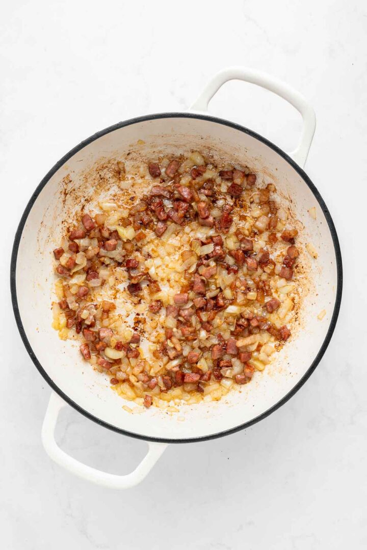 Chopped onions and diced meat sautéing in a white pot on a white surface, creating a savory base that would pair perfectly with comforting Apple Pie Baked Beans.