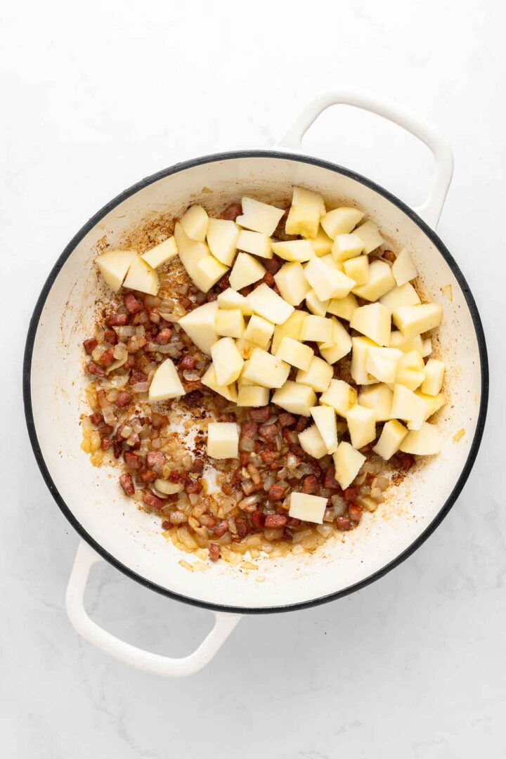 Diced apples, onions, and bacon—just the start of delicious Apple Pie Baked Beans—cooking in a white pot on a white surface.
