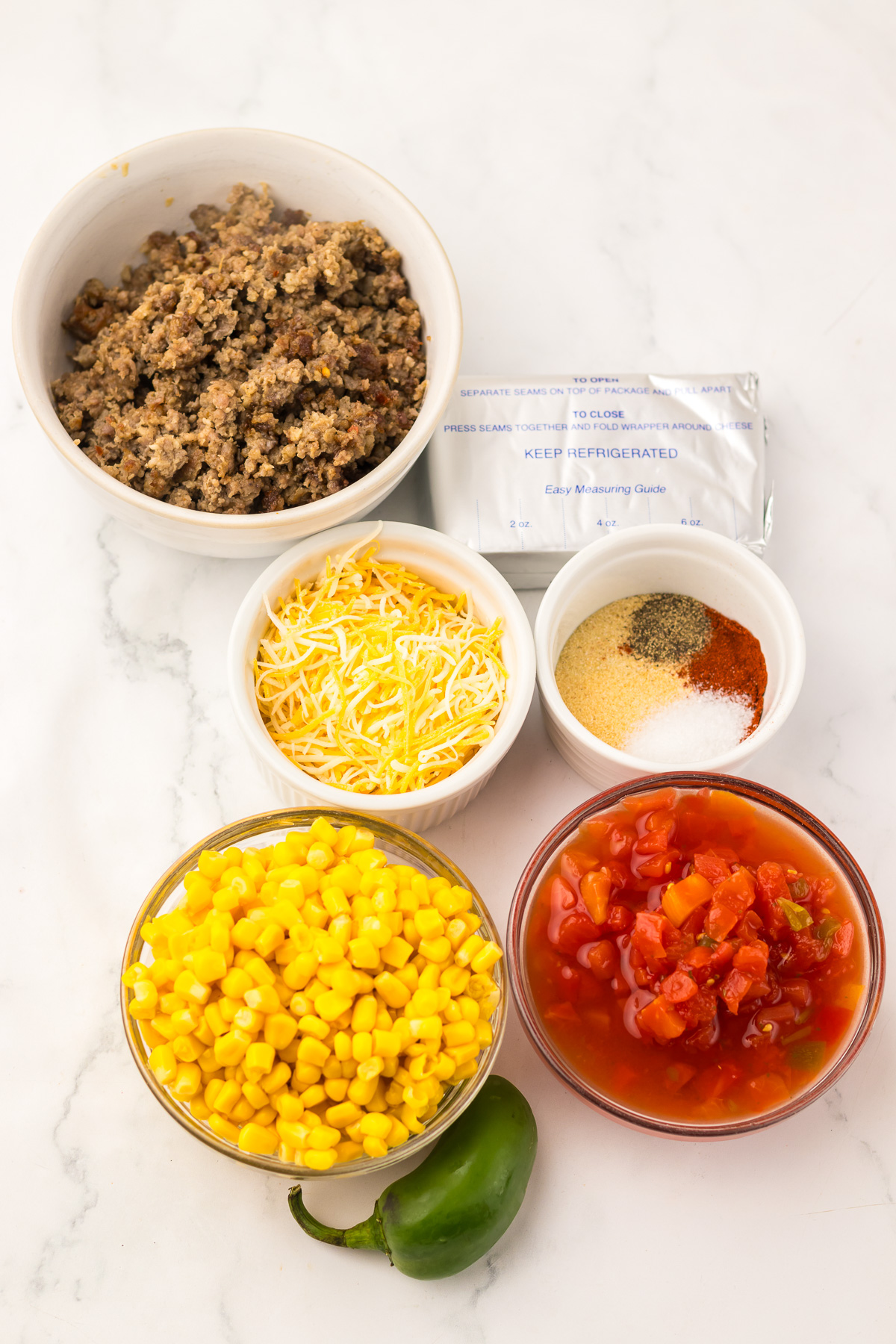 Five bowls with ground meat, shredded cheese, corn, diced tomatoes, seasonings, and a jalape&ntilde;o on a white surface&mdash;perfect ingredients for whipping up a flavorful Slow Cooker Cowboy Dip.