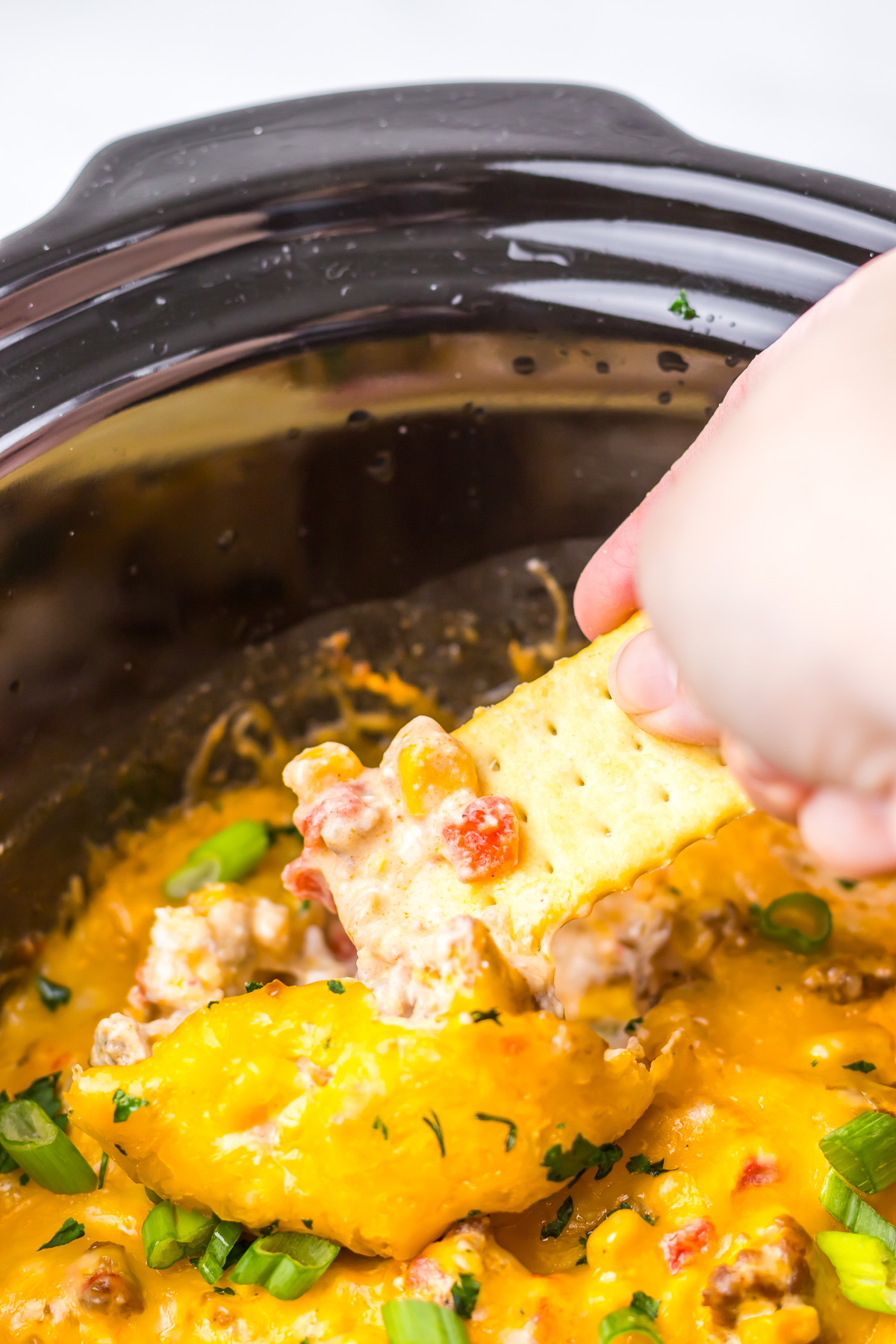 A hand dipping a cracker into cheesy sausage Slow Cooker Cowboy Dip in a slow cooker, topped with green onions.