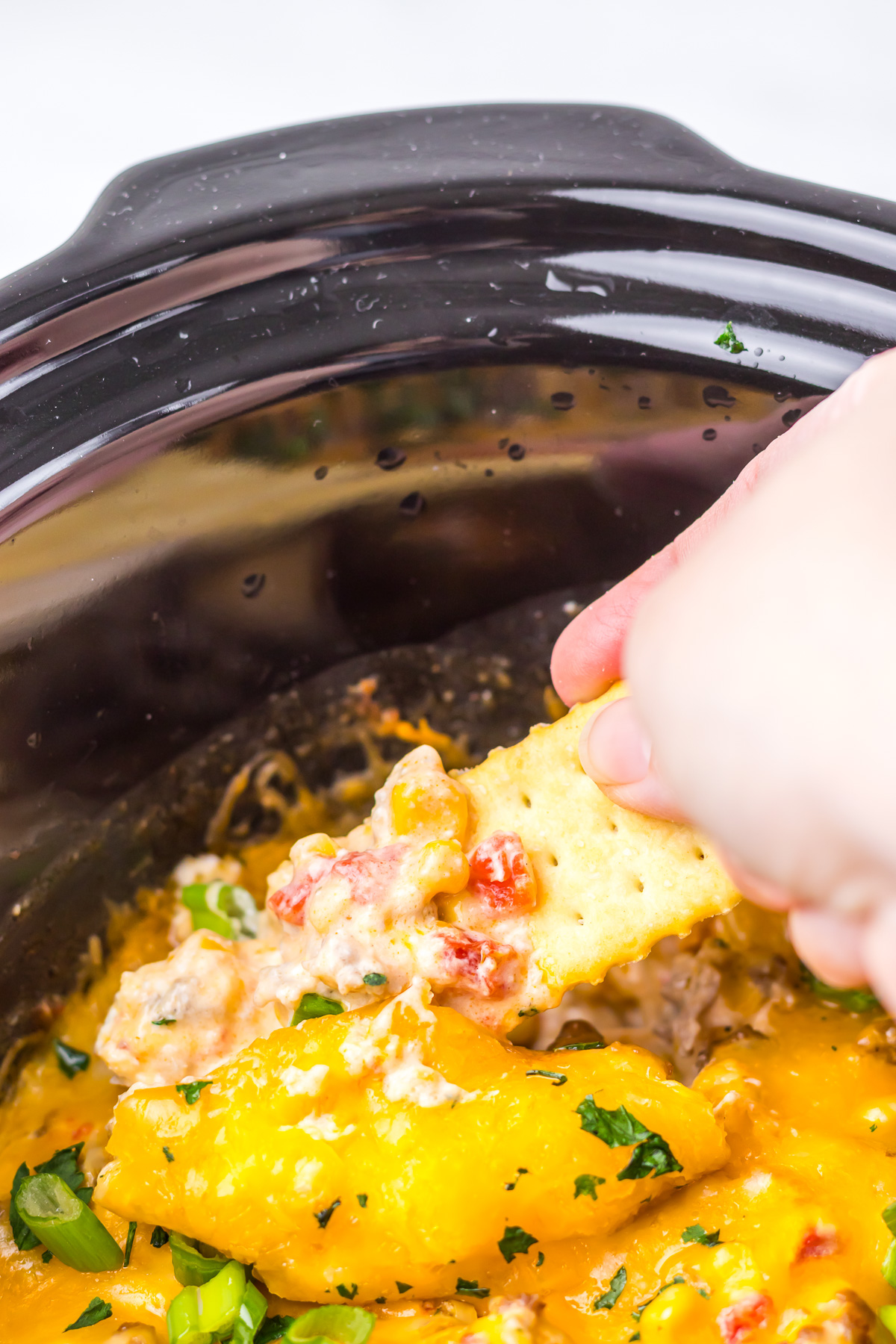 A hand dips a cracker into cheesy Slow Cooker Cowboy Dip with tomatoes in a black slow cooker.