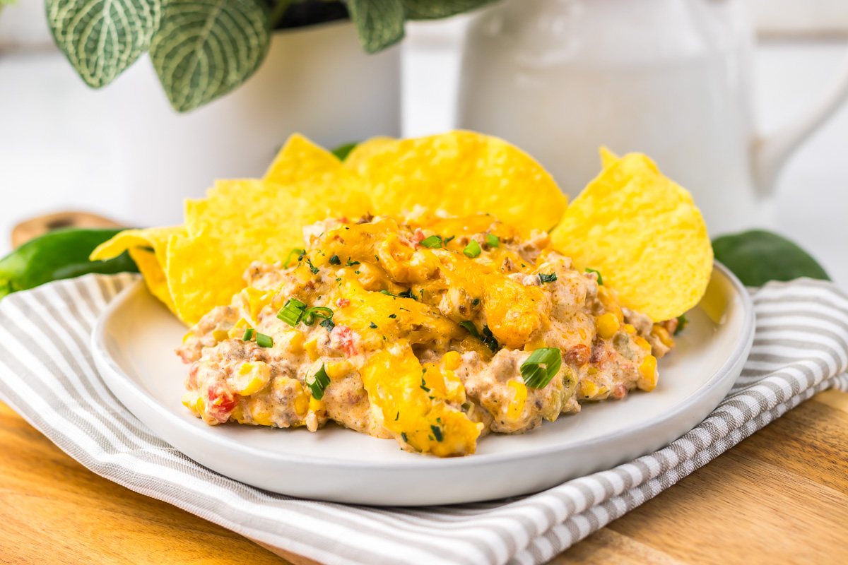 Plate of cheesy Slow Cooker Cowboy Dip with corn, ground meat, and tortilla chips, garnished with chopped herbs on a striped napkin.