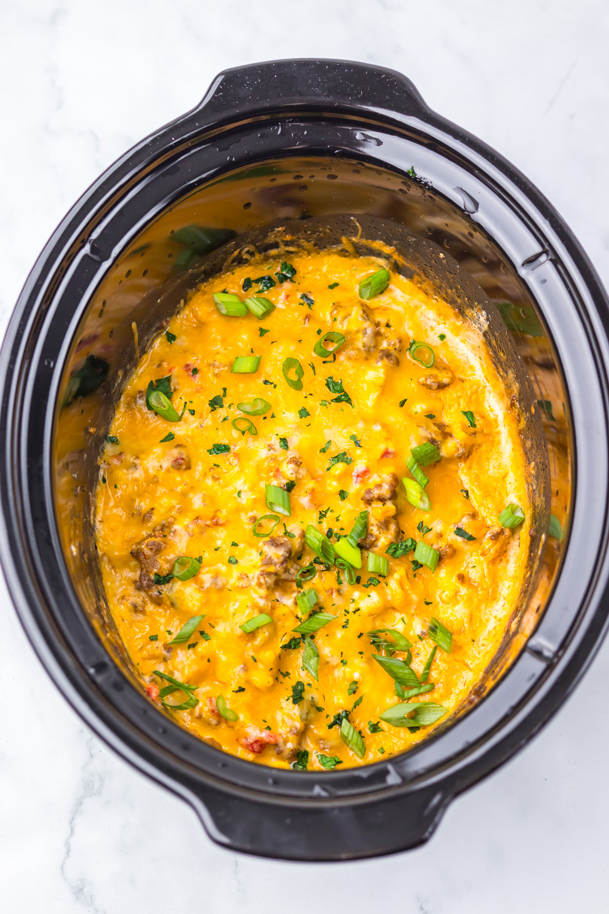 Slow Cooker Cowboy Dip with ground beef and green onions served in a black slow cooker on a white marble surface. This cheesy dip is perfect for gatherings and tailgates, bringing bold flavors and hearty ingredients together for an irresistible appetizer.