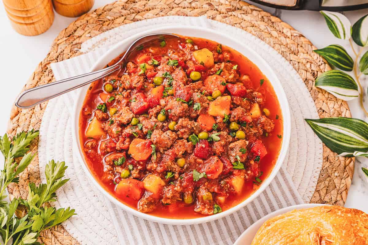 Bowl of hearty Crockpot Poor Man&rsquo;s Stew featuring tender beef with potatoes, peas, and carrots in a rich tomato broth, topped with fresh herbs.