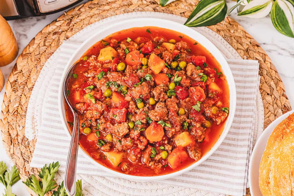 A bowl of Crockpot Poor Man&rsquo;s Stew with peas, carrots, and potatoes sits on a woven placemat with a spoon in the bowl.
