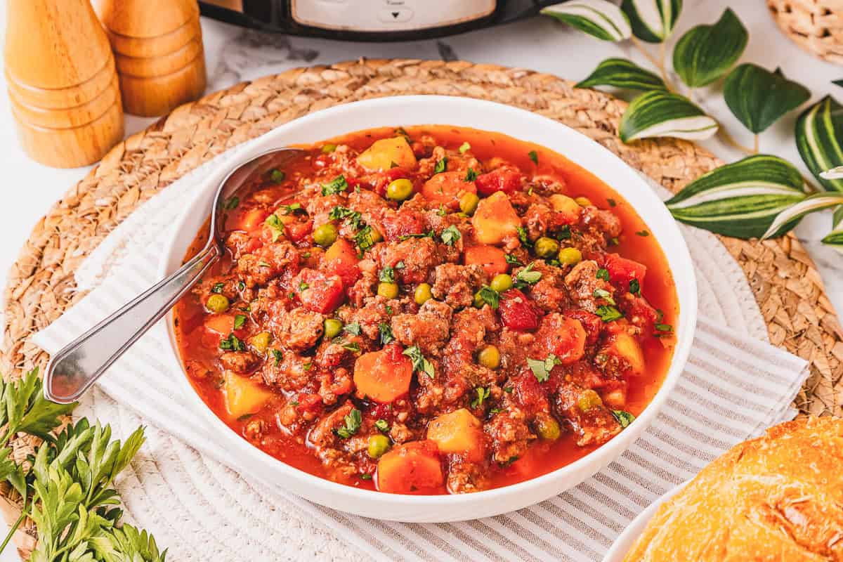 Bowl of Crockpot Poor Man&rsquo;s Stew with beef, vegetables, and tomato sauce, served with a spoon on a striped cloth.