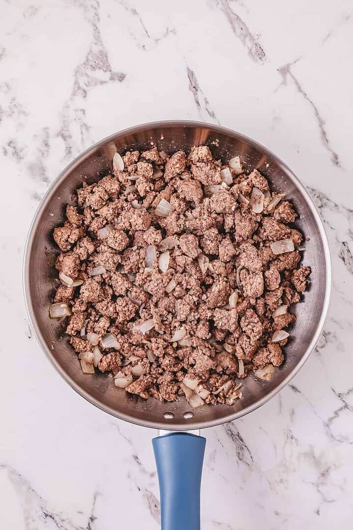 Ground beef and chopped onions cooking in a skillet on a marble countertop, the perfect start for a hearty Crockpot Poor Man&rsquo;s Stew.