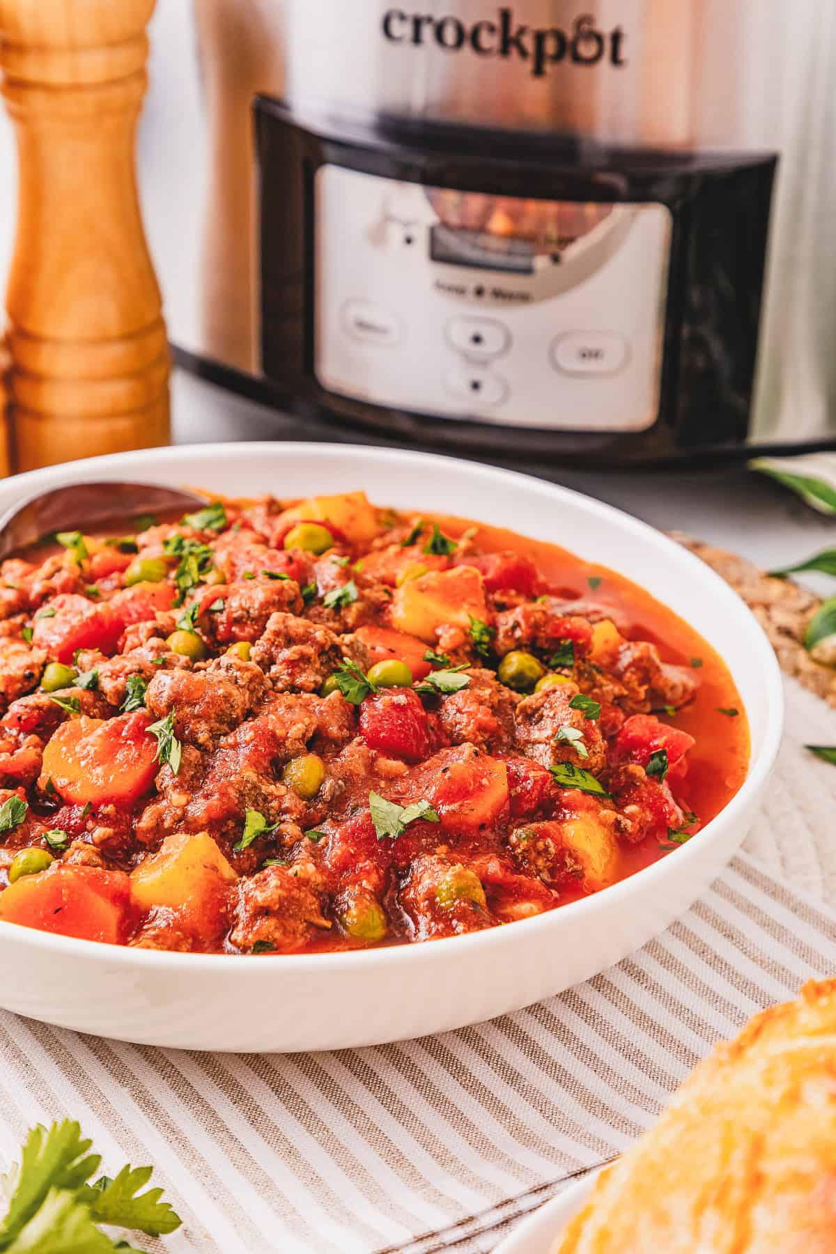 A bowl of hearty Crockpot Poor Man&rsquo;s Stew with vegetables sits in front of a slow cooker on a striped towel.
