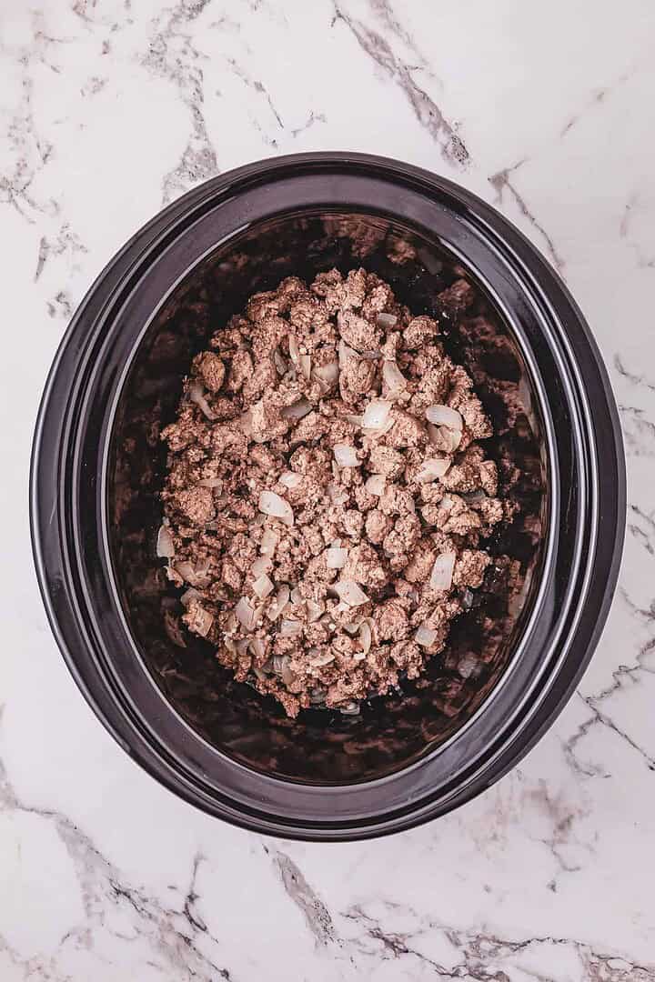 Ground beef and chopped onions in a black slow cooker on a white marble countertop, perfect for starting Crockpot Poor Man&rsquo;s Stew.