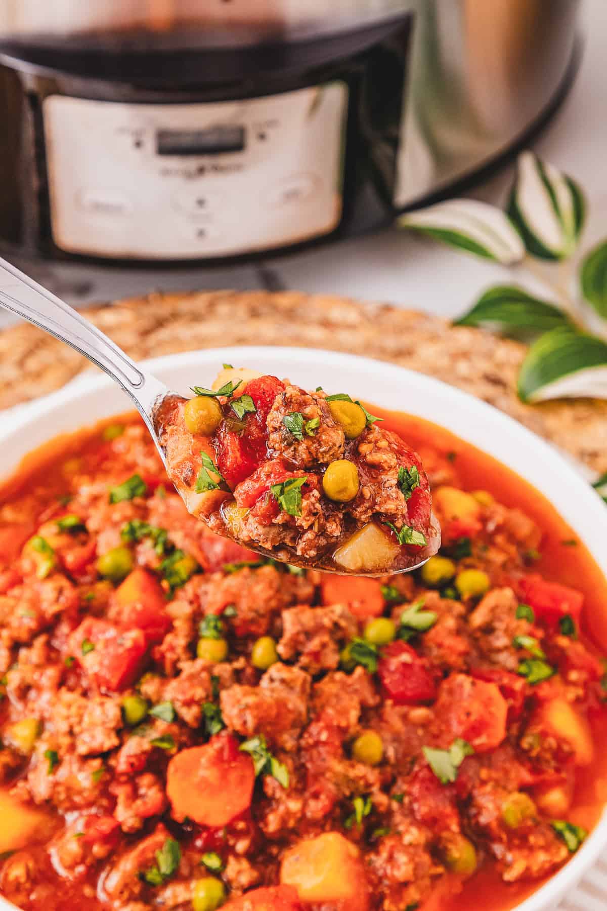 A spoonful of Crockpot Poor Man&rsquo;s Stew with tender beef and vegetables is held above a bowl, with a slow cooker in the background.