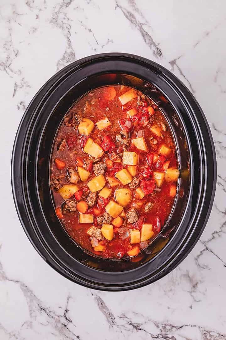 Crockpot Poor Man&rsquo;s Stew simmers in a slow cooker filled with tender beef, potatoes, carrots, and tomatoes in a rich broth on a marble surface.