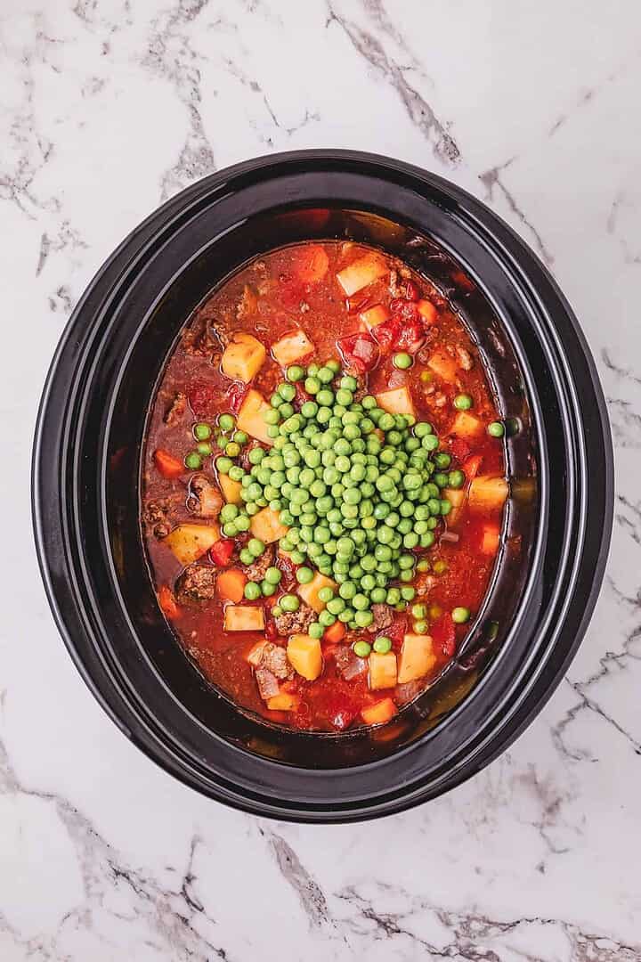 A Crockpot Poor Man&rsquo;s Stew simmers in a slow cooker, filled with hearty vegetable stew and topped with a pile of green peas, all set on a marble countertop.