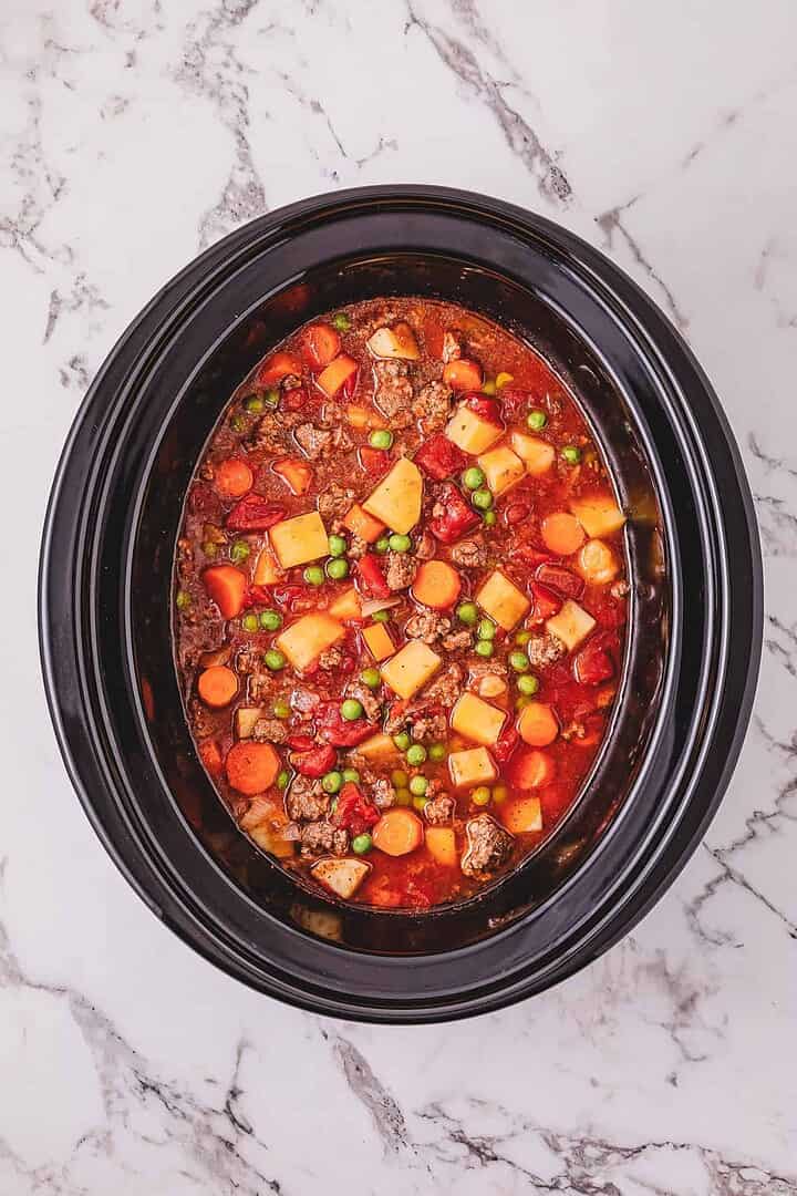 Crockpot Poor Man&rsquo;s Stew with beef, carrots, potatoes, and peas simmering in a black slow cooker on a white marble countertop.