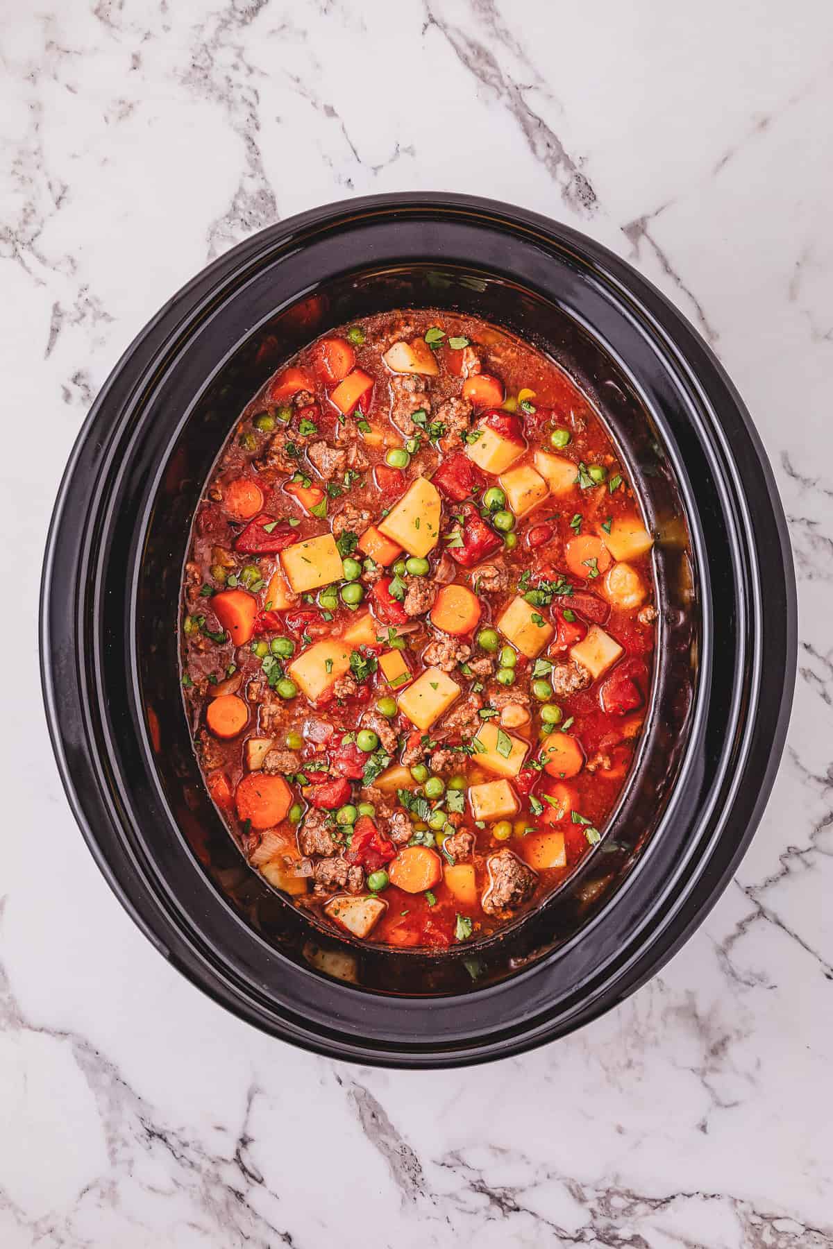 Crockpot Poor Man&rsquo;s Stew simmering in a slow cooker, filled with tender beef, carrots, peas, potatoes, and tomatoes on a marble countertop.