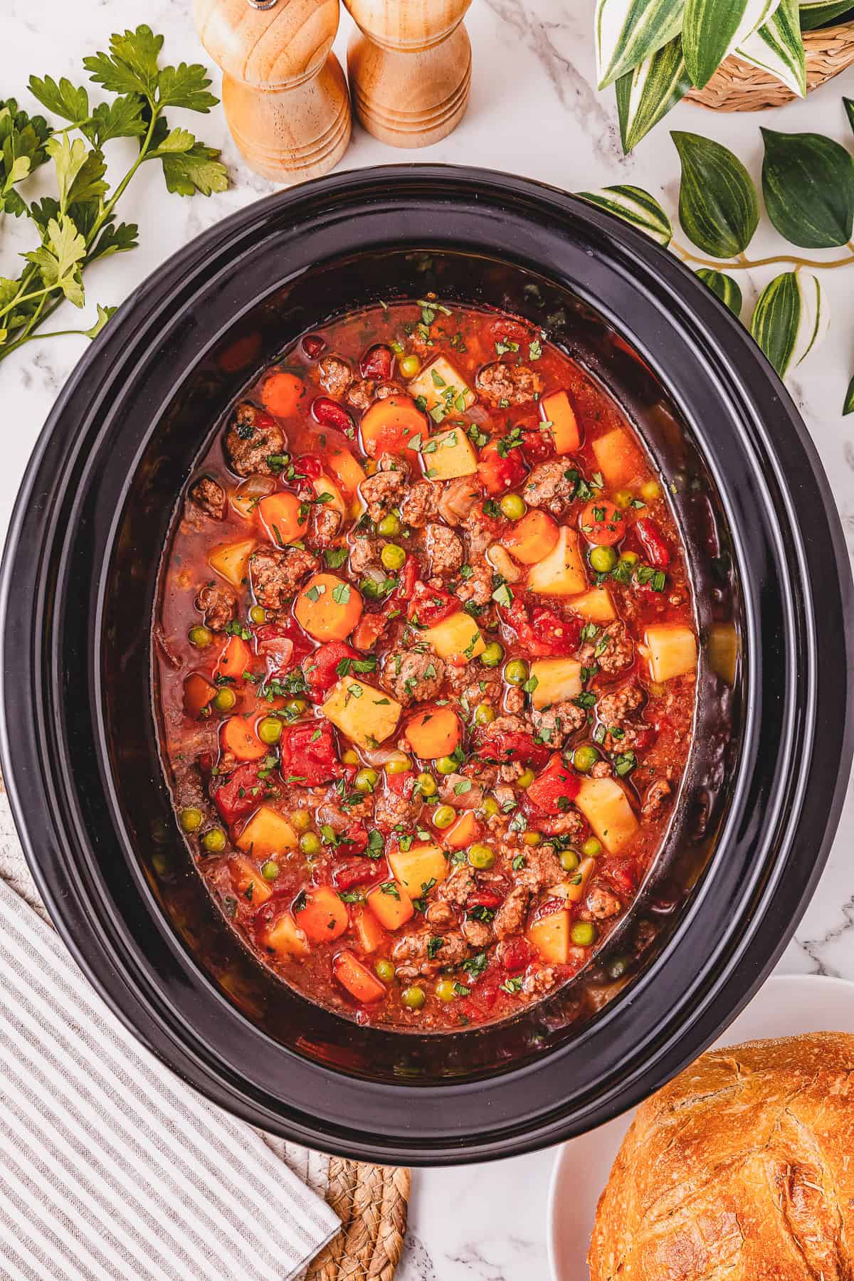 Hearty Crockpot Poor Man&rsquo;s Stew featuring beef and vegetables simmers in a slow cooker, surrounded by rustic bread, fresh herbs, and seasonings on a welcoming table.