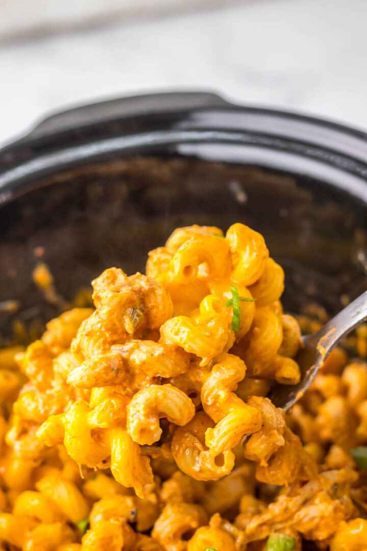 Close-up of cheesy macaroni with ground beef being scooped from a black slow cooker with a metal spoon, reminiscent of the comforting flavors found in slow cooker cheesy chicken enchilada pasta.