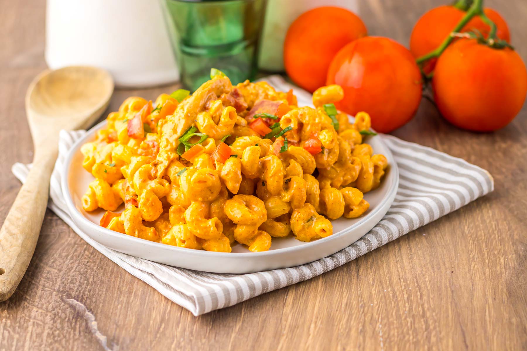 A plate of creamy pasta inspired by slow cooker cheesy chicken enchilada pasta, with diced tomatoes, rests on a striped napkin next to fresh tomatoes and a wooden spoon.