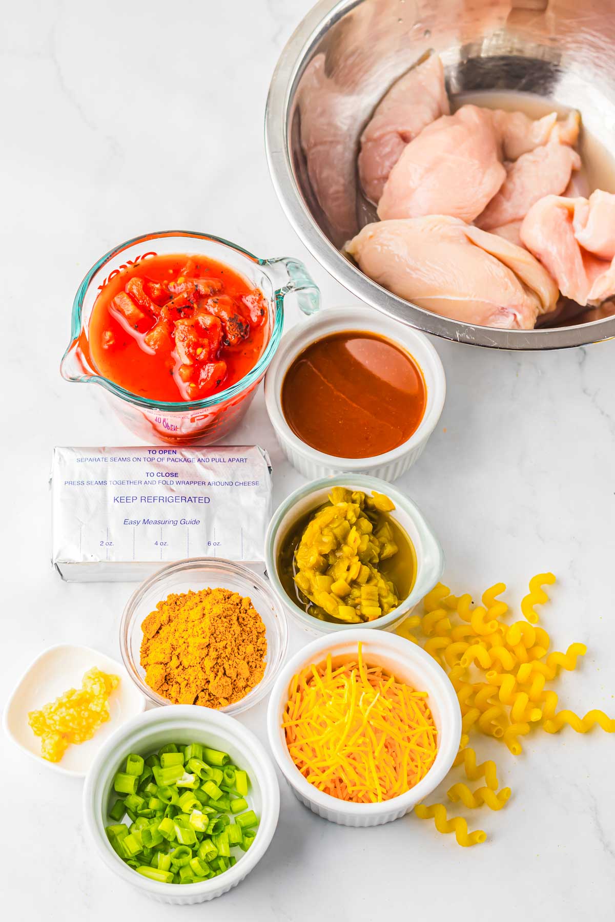 Raw chicken, canned tomatoes, pasta, cheese, spices, and sauces arranged on a white countertop in bowls&mdash;perfectly prepped for making slow cooker cheesy chicken enchilada pasta.