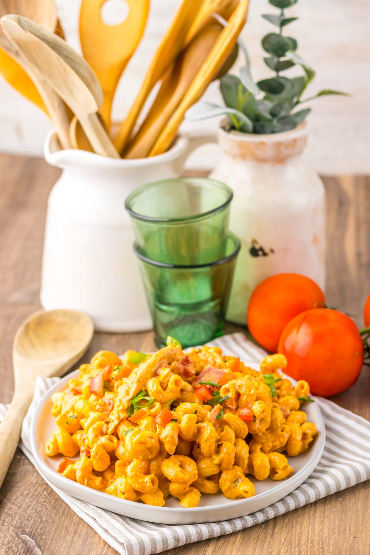 Plate of creamy pasta with vegetables and herbs, reminiscent of slow cooker cheesy chicken enchilada pasta, set on a table with tomatoes, green glasses, and wooden utensils.