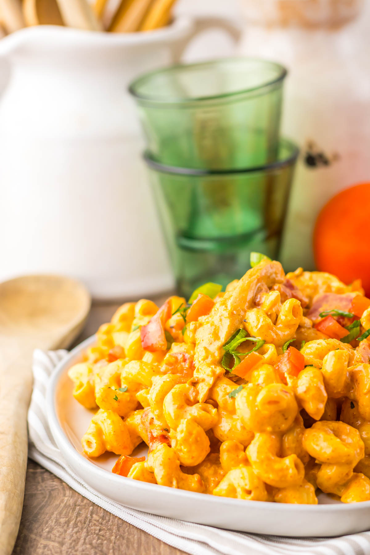 Plate of creamy macaroni salad with vegetables, in front of stacked green glasses and a white pitcher&mdash;a classic side dish perfect for pairing with hearty mains like slow cooker cheesy chicken enchilada pasta.
