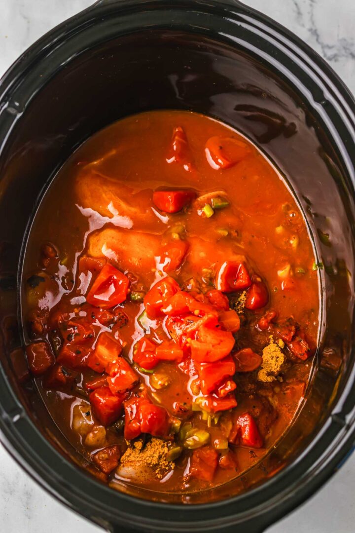 Chicken breasts in a slow cooker with diced tomatoes, sauce, and spices, coming together to create a delicious base for slow cooker cheesy chicken enchilada pasta.