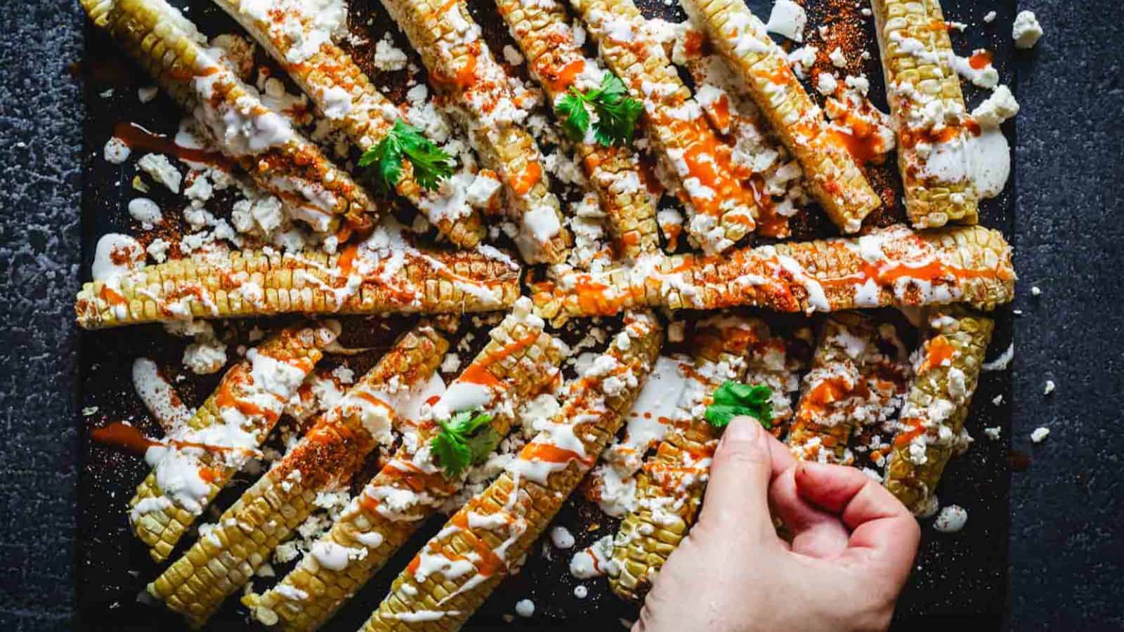 Hand garnishing sliced grilled corn topped with cheese, crema, and sauce on a dark surface.