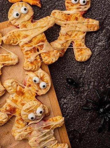 Pastry mummy hand pies with candy eyes on a wooden cutting board next to a bowl of red jam.