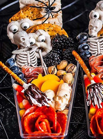 Three Halloween snack cups with crackers, cheese, meats, nuts, fruit, and candy skeletons, spiders, and eyes.