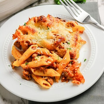 A serving of sausage pasta bake with meat sauce and melted cheese on a white plate with a fork.